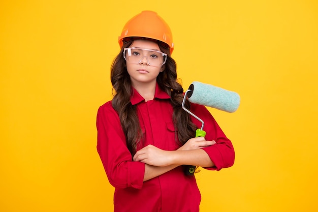 Little builder expert in helmet on construction site teen girl\
painter with painting brush tool or paint roller child on repairing\
work renovation concept