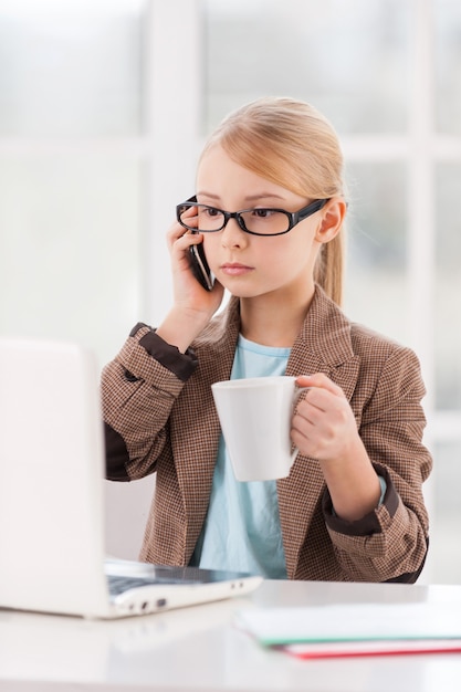 Piccola imprenditrice. fiduciosa bambina con gli occhiali e l'abbigliamento formale che parla al telefono mentre è seduta al tavolo e guarda il laptop