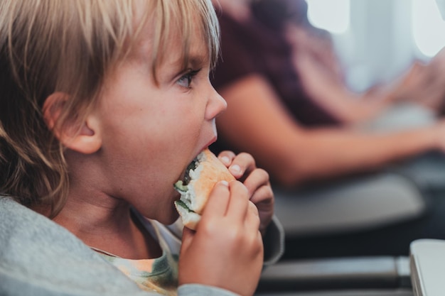 Little candid kid boy five years old eats burger or sandwich food sitting in airplane seat on flight traveling from airport children take a bite child in air plane eating lunch or dinner meal