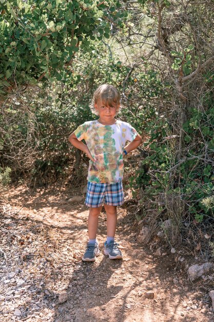 Little candid tired kid boy traveler and hiker of five years\
old in shorts and a tshirt travels and hiking a mountain path among\
forest green plants in the mediterranean during her summer\
vacation