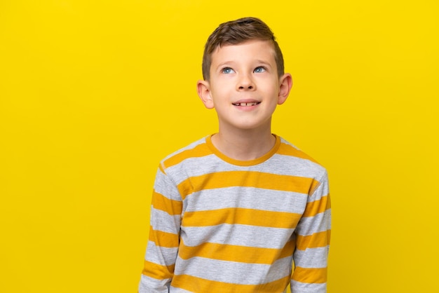 Little caucasian boy isolated on yellow background thinking an idea while looking up