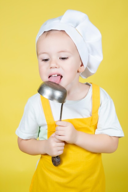 シェフを演じる小さな白人の少年、エプロンとシェフの帽子の少年は黄色の背景に取鍋をなめる