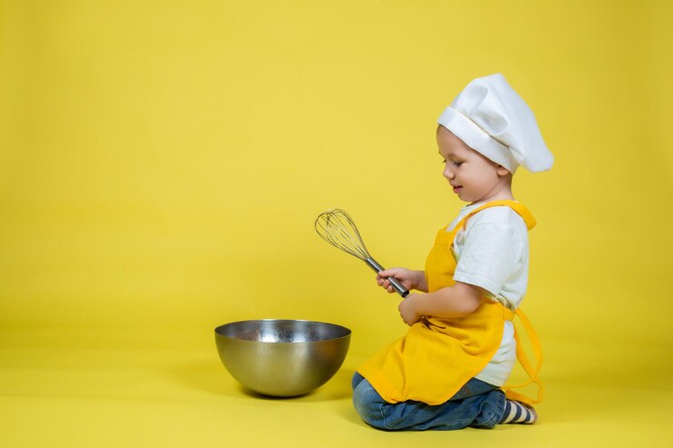 Photo little caucasian boy playing chef, boy in apron and chef's hat sitting on the floor with a bowl and whisk on yellow background