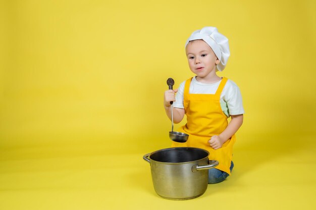 작은 백인 소년 요리사, 앞치마를 입은 소년, 노란색 배경에 냄비와 국자로 바닥에 앉아있는 요리사 모자
