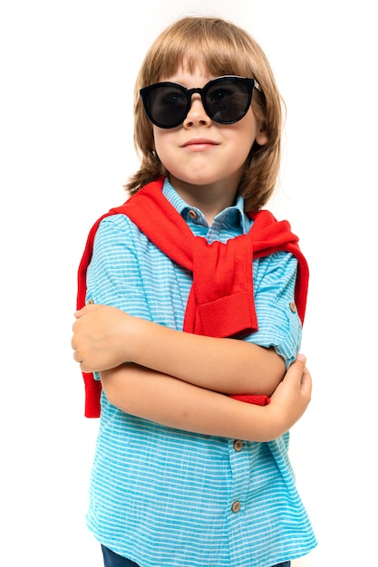 Little caucasian boy sits on a chair with red sweatshot around his neck and sunglasses isolated on white surface