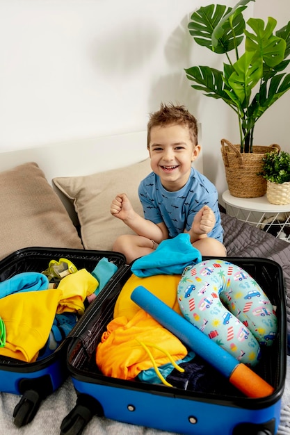 Photo little caucasian boy with blue shirt ready for vacation. happy child packs clothes into a suitcase for travel. tourist, joy of holiday. kid at home, preparing for flying. modern and cozy interior.