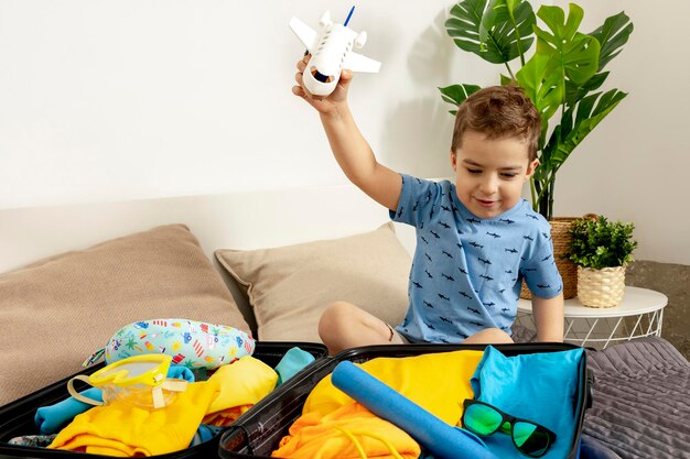 Little caucasian boy with blue shirt ready for vacation Happy child packs clothes into a suitcase for travel Tourist joy of holiday Kid at home preparing for flying Modern and cozy interior