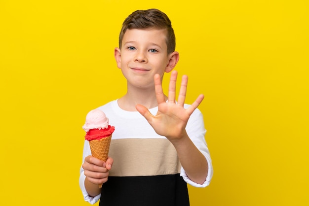 Little caucasian boy with a cornet ice cream isolated on yellow background counting five with fingers