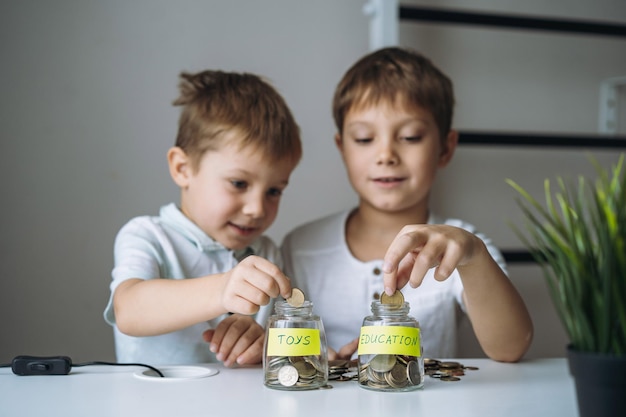 Маленькие кавказские мальчики кладут монету в стеклянную бутылку за столом. Ребенок экономит деньги на образовании.