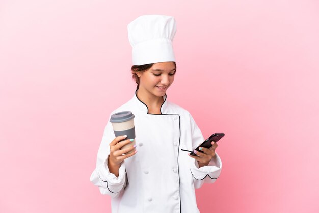 Little caucasian chef girl isolated on pink background holding coffee to take away and a mobile