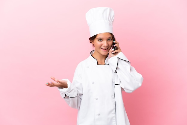 Little caucasian chef girl isolated on pink background keeping a conversation with the mobile phone with someone