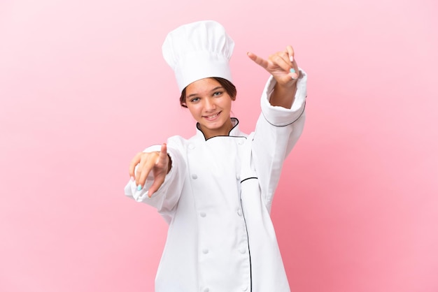 Little caucasian chef girl isolated on pink background points finger at you while smiling