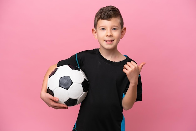 Little caucasian football player boy isolated on pink background pointing to the side to present a product