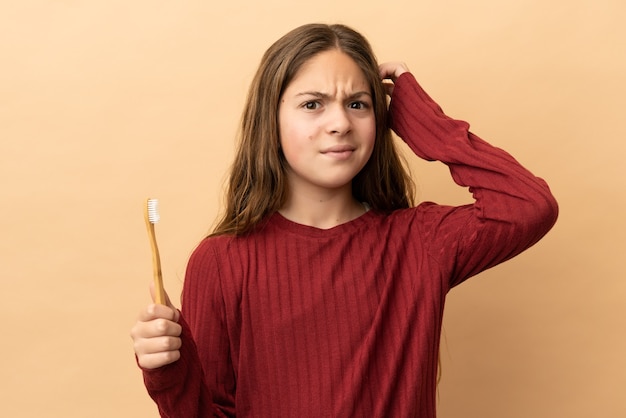 Little caucasian girl brushing her teeth isolated on beige background having doubts