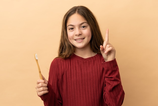 Little caucasian girl brushing her teeth isolated on beige background showing and lifting a finger in sign of the best