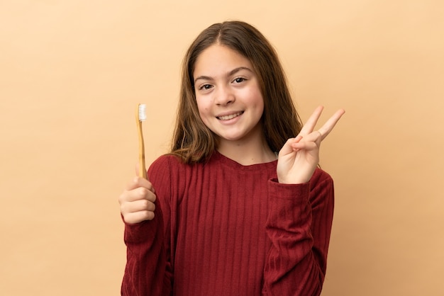 Little caucasian girl brushing her teeth isolated on beige background smiling and showing victory sign