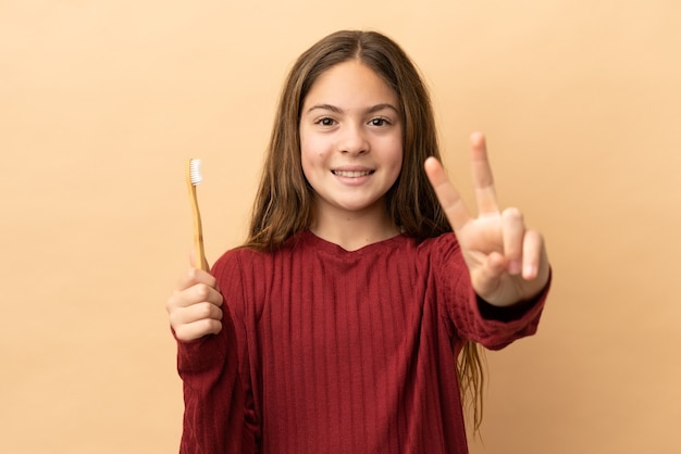 Little caucasian girl brushing her teeth isolated on beige background smiling and showing victory sign