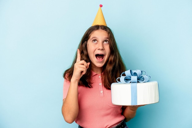 Photo little caucasian girl celebrating her birthday holding a cake isolated on blue background pointing upside with opened mouth.