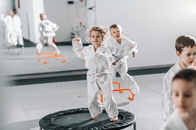 Little Caucasian girl in dobok jumping on trampoline and warming up for taekwondo training.