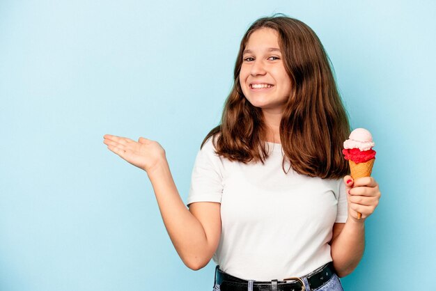 Little caucasian girl eating an ice cream isolated on blue background showing a copy space on a palm and holding another hand on waist.