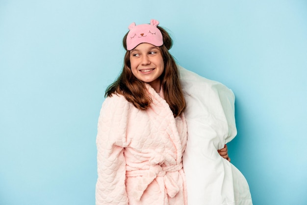 Photo little caucasian girl going to sleep isolated on blue background looks aside smiling, cheerful and pleasant.