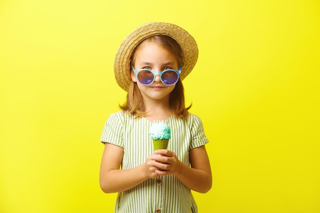 Little caucasian girl in green dress and straw hat eating beautiful ice cream wears blue sunglasses has a good mood stands on yellow isolated background