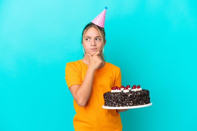 疑問を持っている青い背景に分離されたバースデーケーキを保持している小さな白人の女の子