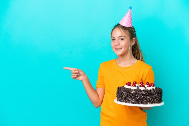 Little caucasian girl holding birthday cake isolated on blue background pointing finger to the side