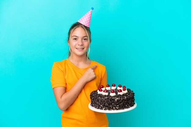 Little caucasian girl holding birthday cake isolated on blue\
background pointing to the side to present a product