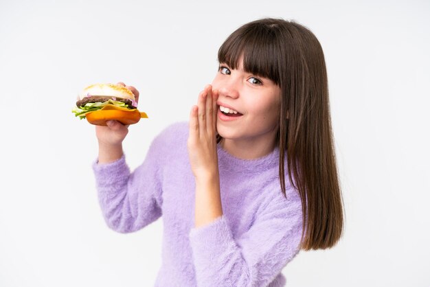 Маленькая кавказская девочка держит гамбургер на изолированном фоне и что-то шепчет