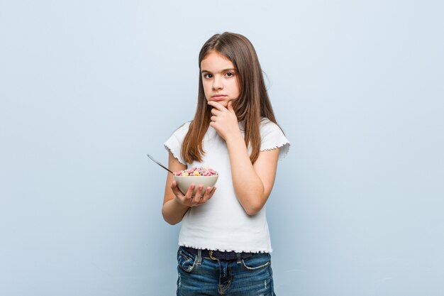 Little caucasian girl holding a cereal bowl looking sideways with doubtful and skeptical expression.