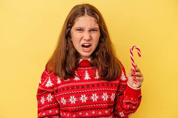 Piccola ragazza caucasica che tiene il bastone di natale isolato su sfondo giallo che urla molto arrabbiato e aggressivo.