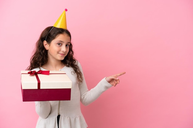 Little caucasian girl holding a gift isolated on pink background surprised and pointing finger to the side
