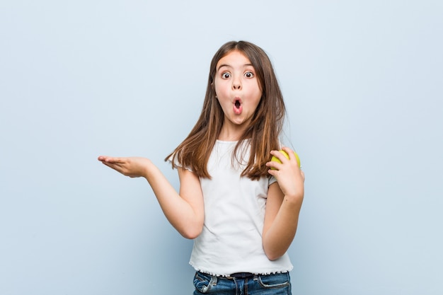 Little caucasian girl holding a green apple impressed holding copy space on palm.