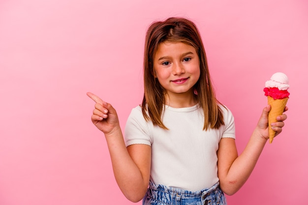 Little caucasian girl holding ice cream isolated on pink smiling and pointing aside, showing something at blank space.