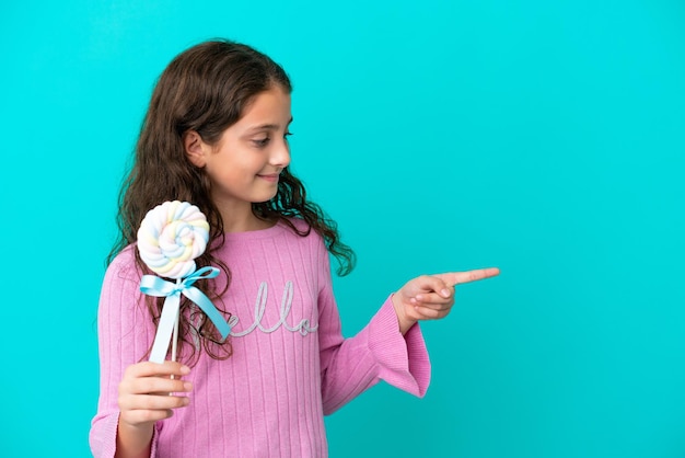 Little caucasian girl holding a lollipop isolated on blue background pointing to the side to present a product