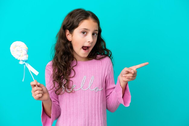 Photo little caucasian girl holding a lollipop isolated on blue background surprised and pointing side