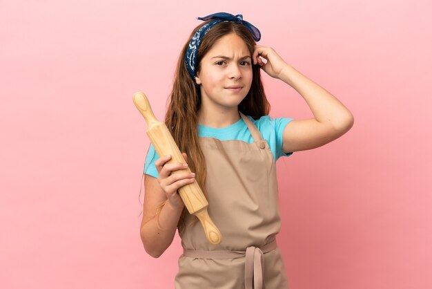 Little caucasian girl holding a rolling pin isolated on pink background having doubts