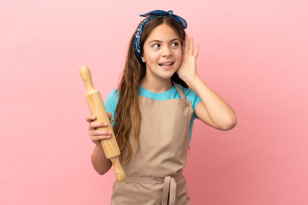 Little caucasian girl holding a rolling pin isolated on pink background listening to something by putting hand on the ear