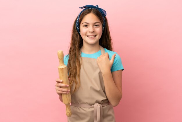 Little caucasian girl holding a rolling pin isolated on pink background pointing to the side to present a product