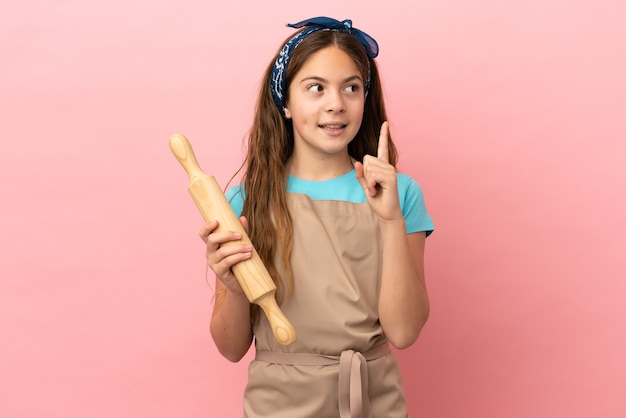 Little caucasian girl holding a rolling pin isolated on pink background thinking an idea pointing the finger up