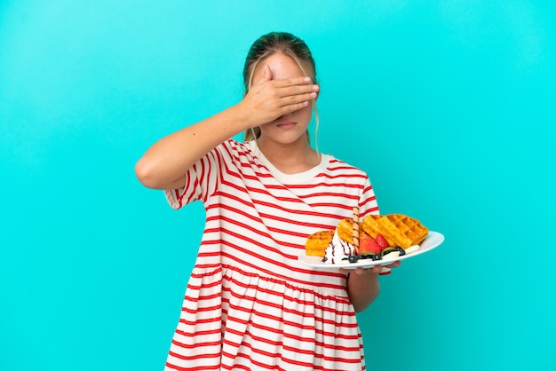 Маленькая кавказская девочка держит вафли на синем фоне, закрывая глаза руками. Не хочу видеть что-то