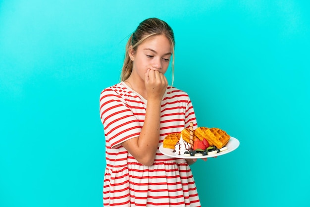 Little caucasian girl holding waffles isolated on blue background having doubts