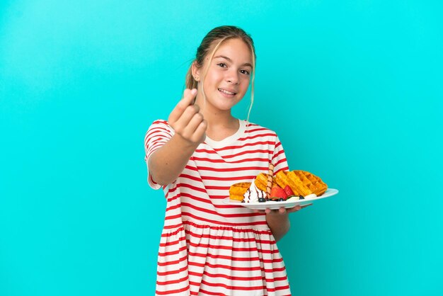 Little caucasian girl holding waffles isolated on blue background making money gesture