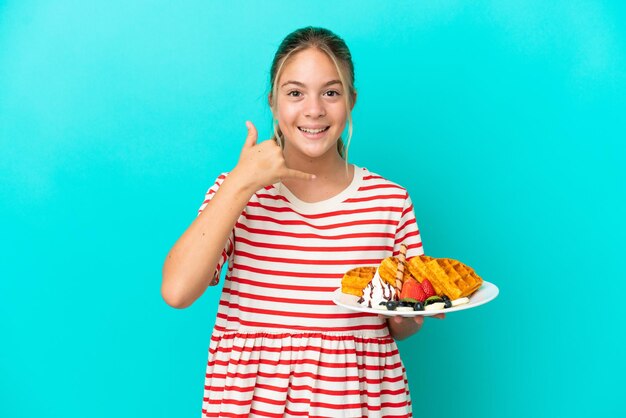 Little caucasian girl holding waffles isolated on blue background making phone gesture Call me back sign