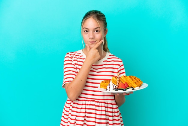 Little caucasian girl holding waffles isolated on blue background thinking