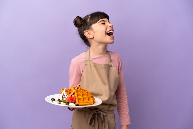 Little caucasian girl holding waffles isolated on purple wall laughing