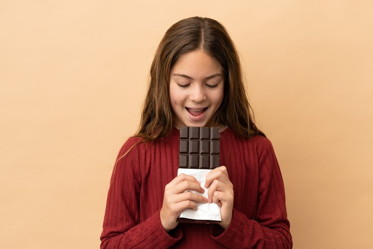 Photo little caucasian girl isolated on beige background eating a chocolate tablet