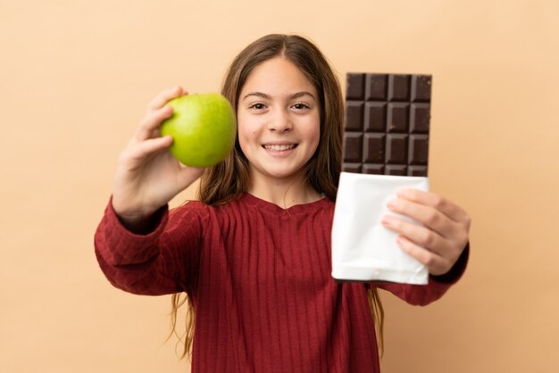 Little caucasian girl isolated on beige background taking a chocolate tablet in one hand and an apple in the other