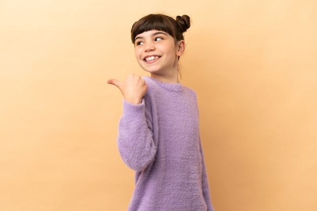 Little caucasian girl isolated on beige pointing to the side to present a product